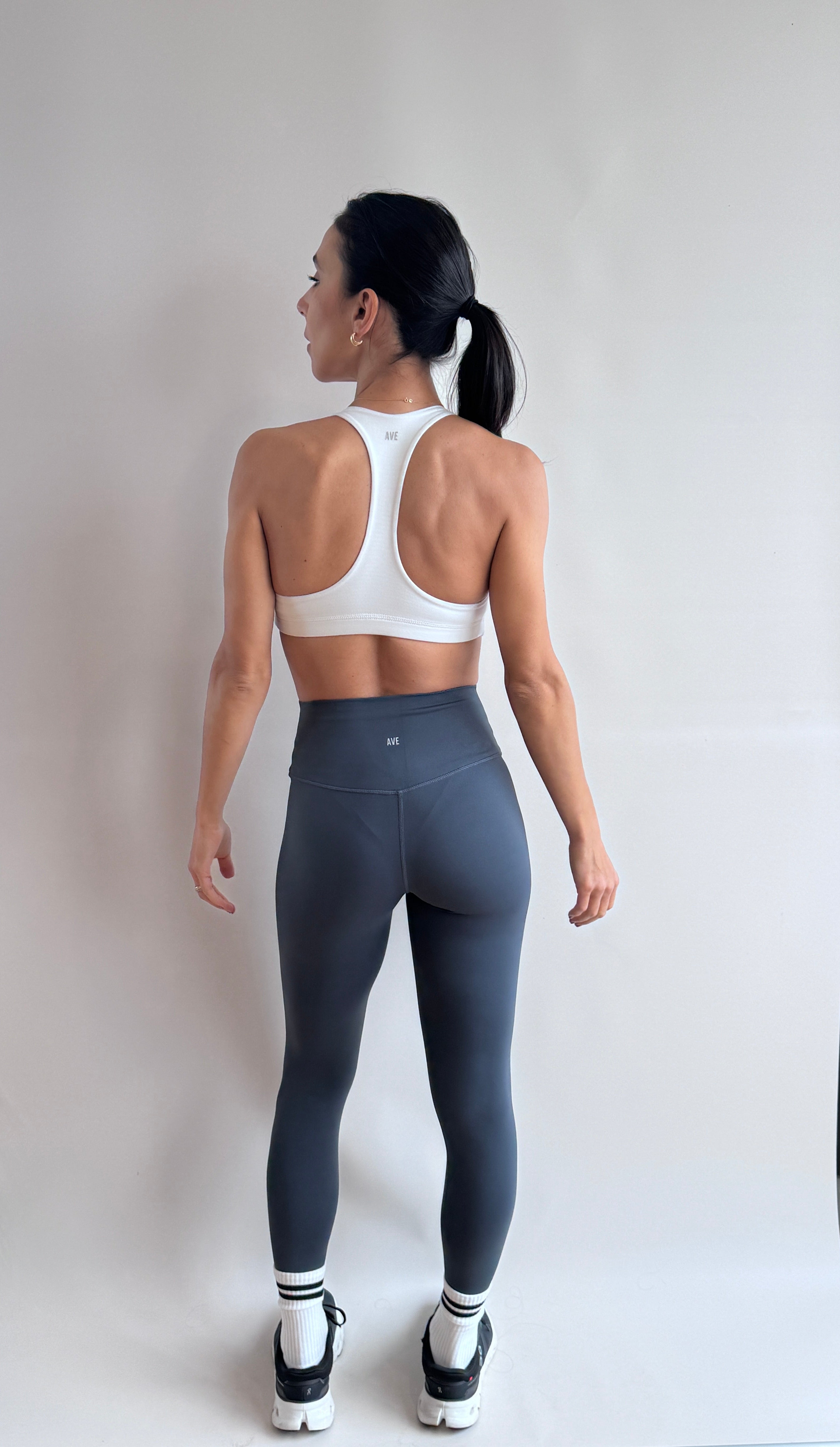 A woman stands with her back to the camera wearing a white sports bra and dark gray high-waisted leggings, standing in front of a white studio backdrop.