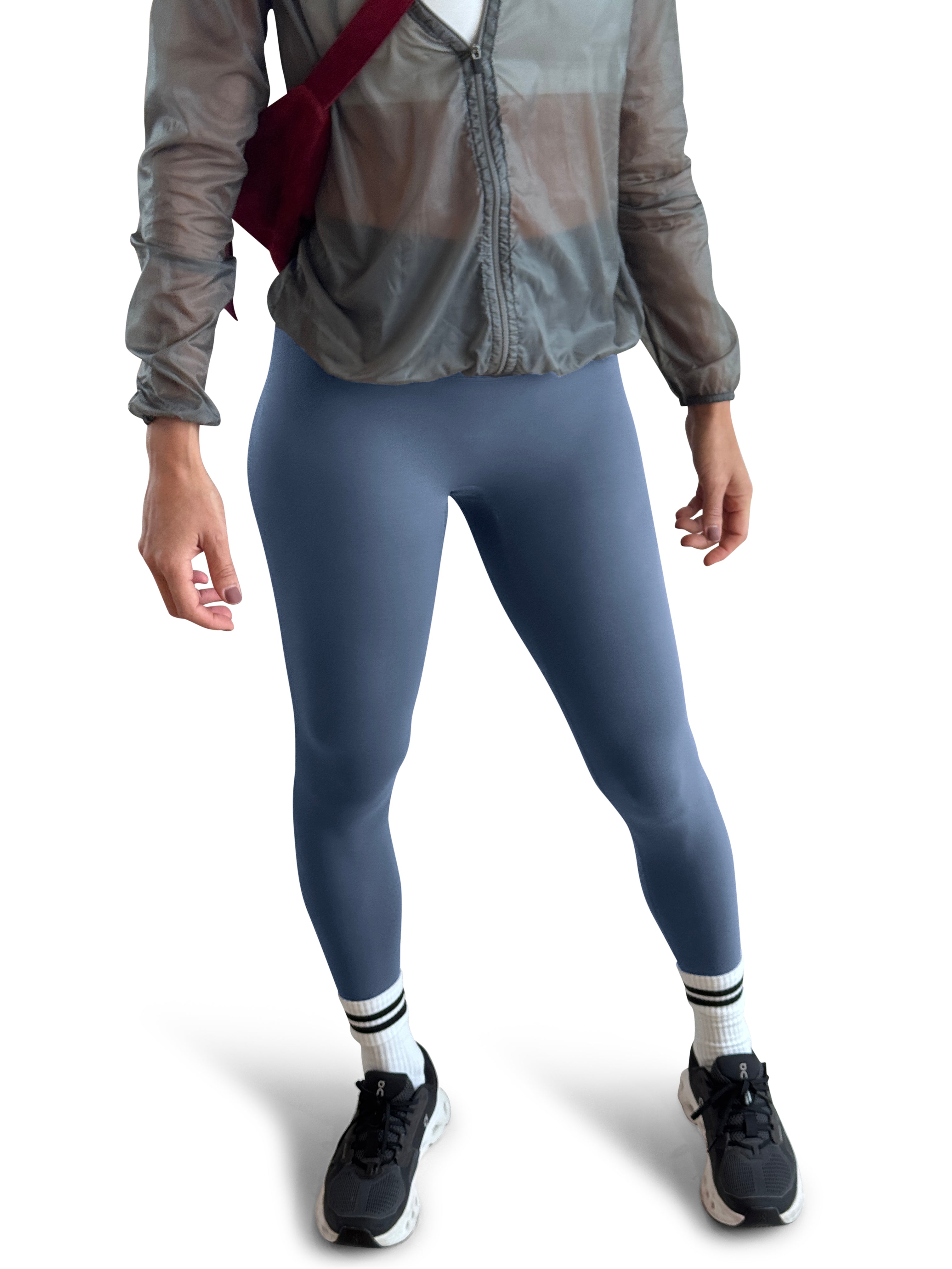 A model wearing slate blue leggings, a sheer gray zip-up jacket, white socks, and black running shoes, carrying a dark red shoulder bag against a white background.