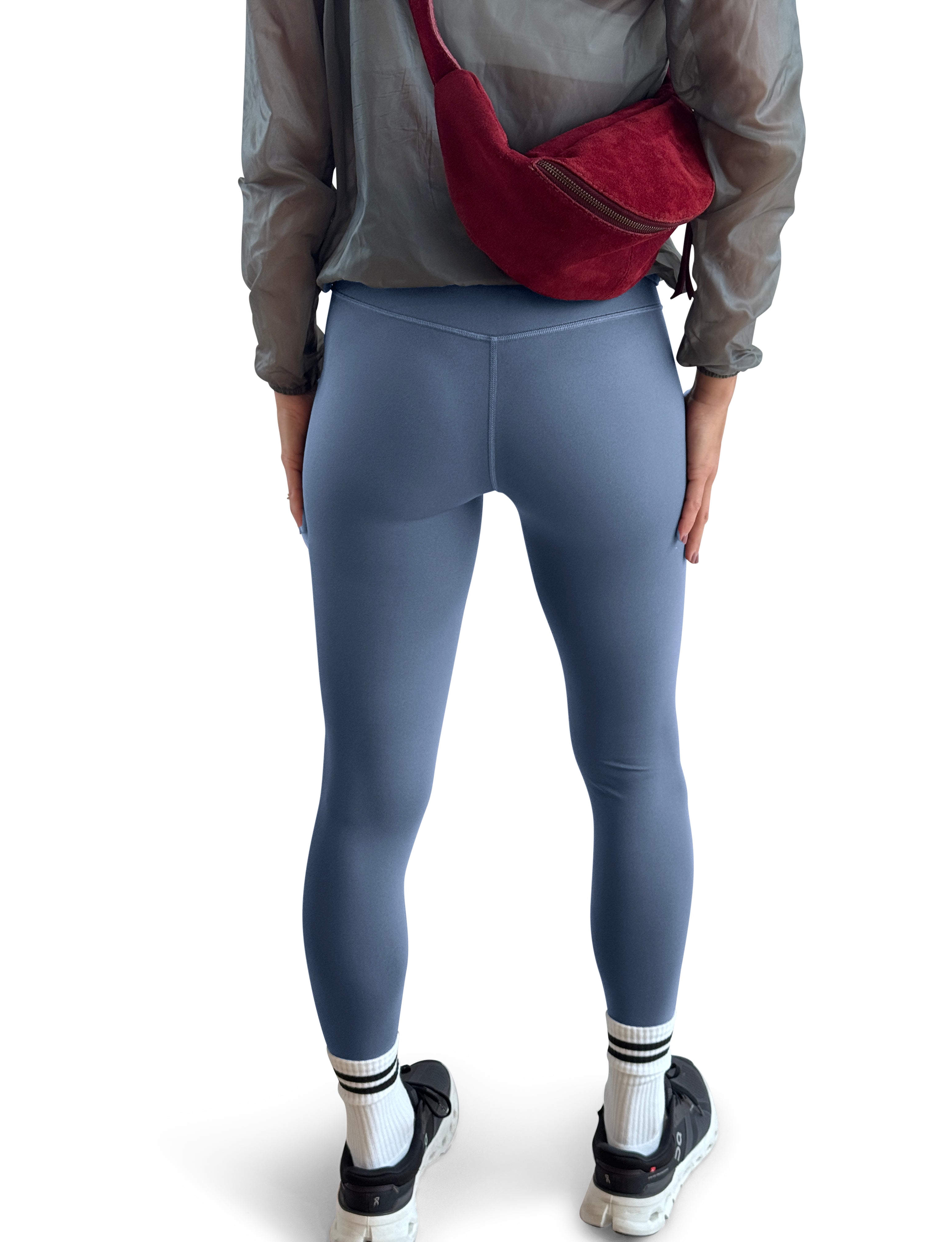 A person shown from the back wearing high-waisted slate blue leggings, a sheer gray long-sleeve top, white socks, and black sneakers, with a red crossbody bag worn across the upper back, against a plain white background.