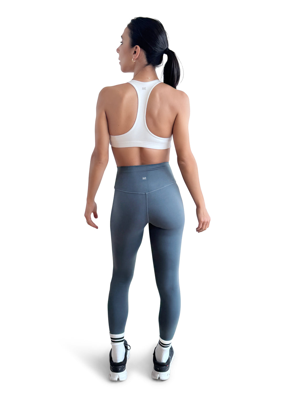A woman is shown from behind wearing a white sports bra and slate blue high-waisted leggings, standing against a solid white background.