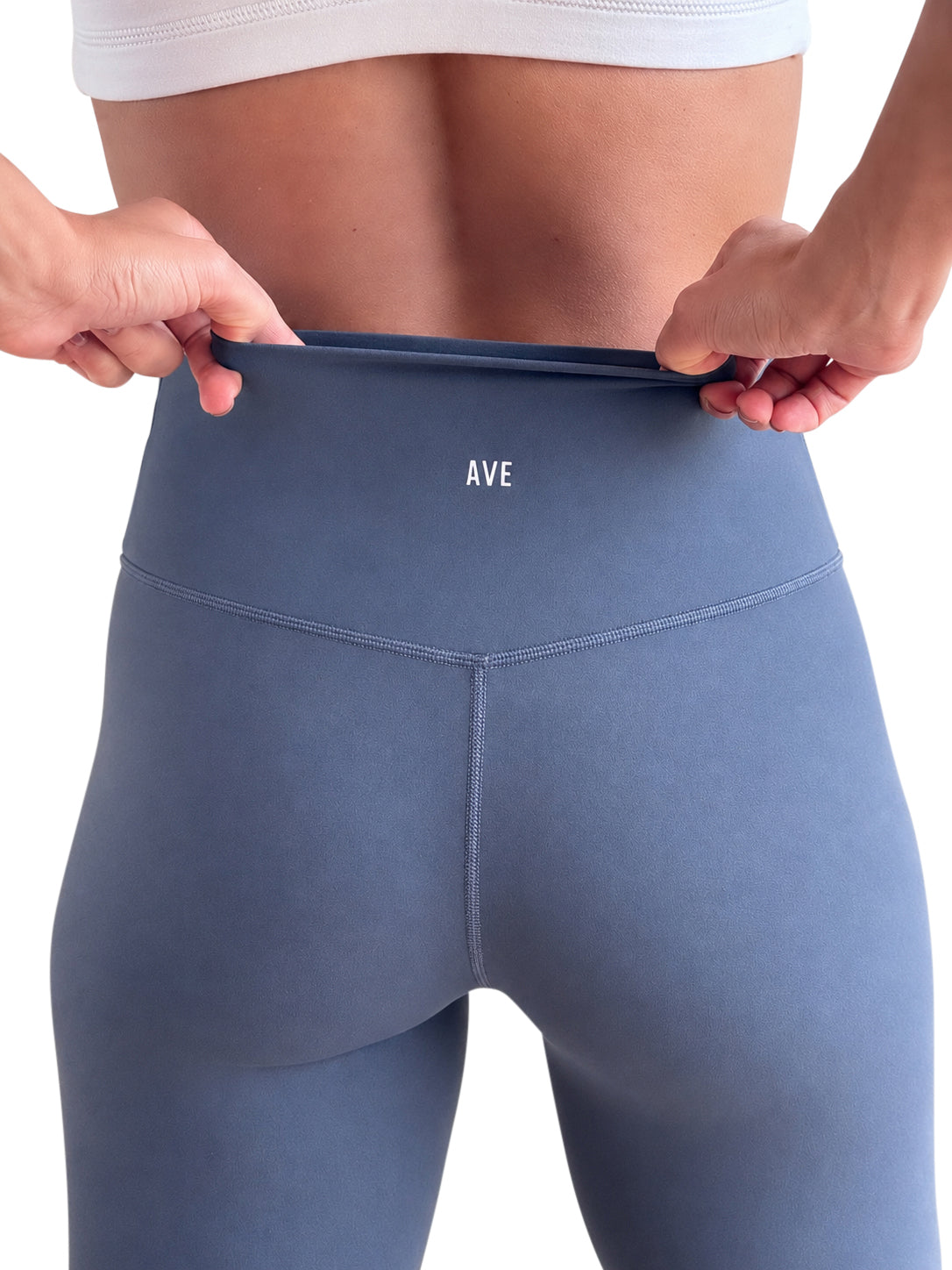 Close-up of a person wearing high-waisted blue-gray leggings, pulling the waistband outward with both hands. The brand name 'AVE' is printed in white on the back waistband.